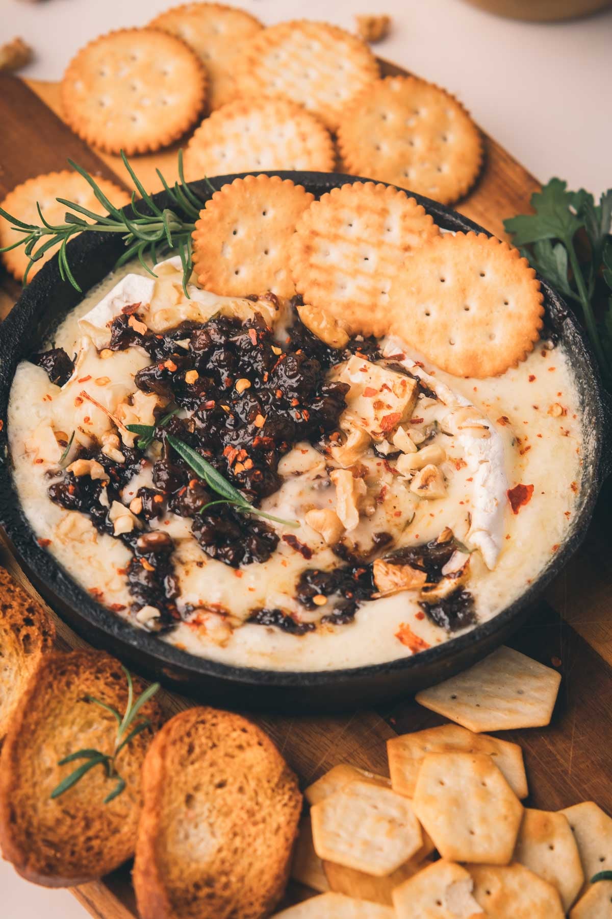 A bowl of baked brie topped with nuts and bacon jam, surrounded by round crackers and toasted bread on a wooden board.