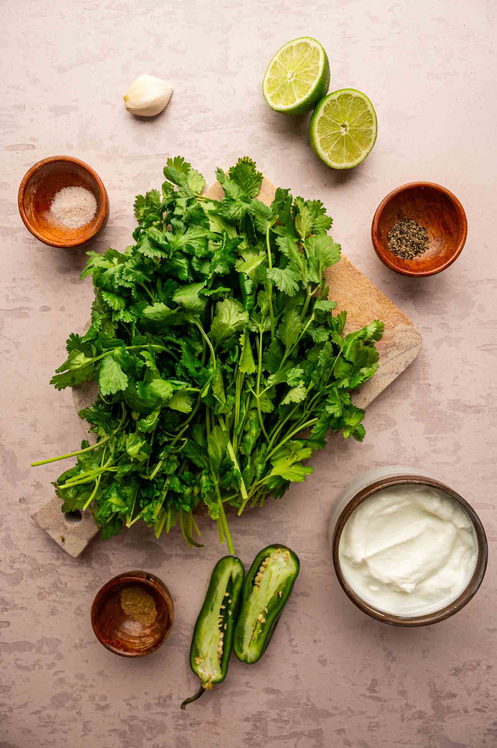 Fresh cilantro on a cutting board surrounded by a halved lime, halved jalapeño, garlic clove, small bowls of spices, and a bowl of yogurt on a light surface.