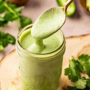 A spoon scoops creamy green sauce from a glass jar, surrounded by fresh cilantro, sliced jalapeño, lime, and a bowl of pepper.