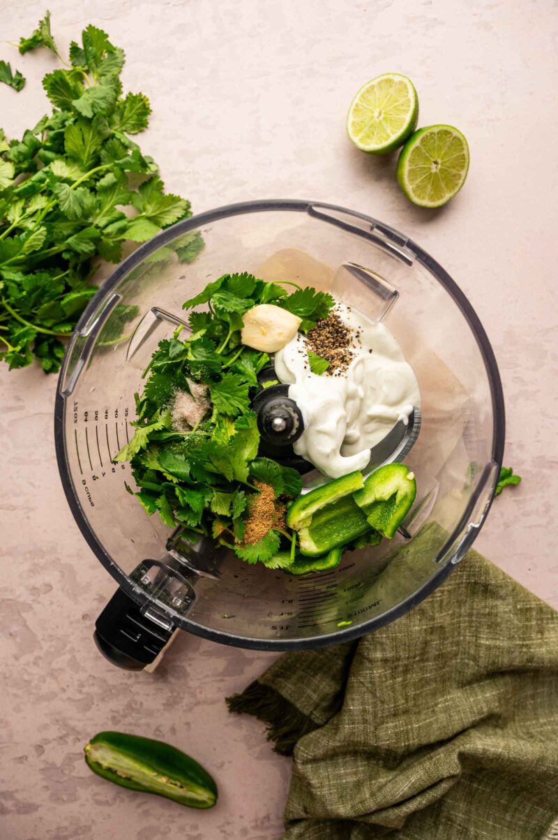 A food processor bowl containing cilantro, sliced jalapeño, garlic, Greek yogurt, spices, salt, and pepper, with fresh cilantro and halved limes nearby.