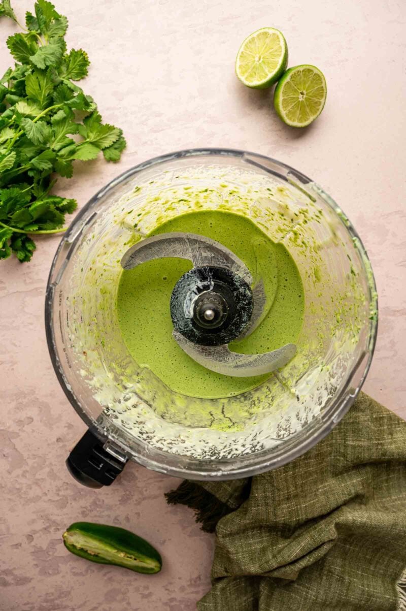 A food processor that's just pureed cilantro sauce sits on a countertop with cilantro, a halved lime, a jalapeño, and a green cloth nearby.