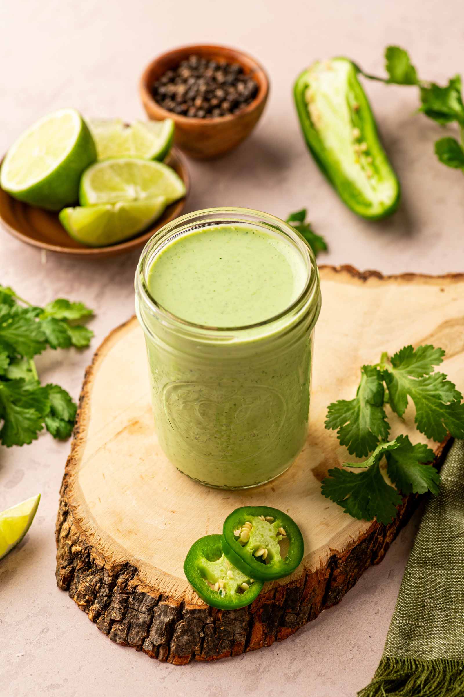 A jar of green cilantro sauce sits on a wooden board, surrounded by lime wedges, cilantro, jalapeño slices, a halved jalapeño, and a small bowl of black pepper.