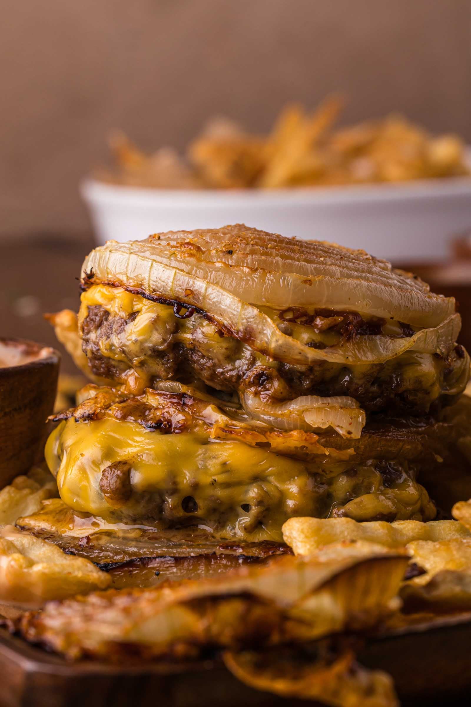 A close-up of a stacked Flying dutchman burger with melted cheese and baked onions, with potato chips in the background.
