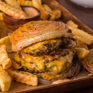 A Flying dutchman burger with grilled onions as buns, melted cheese, and crinkle-cut fries on a tray, with animal sauce on the side.