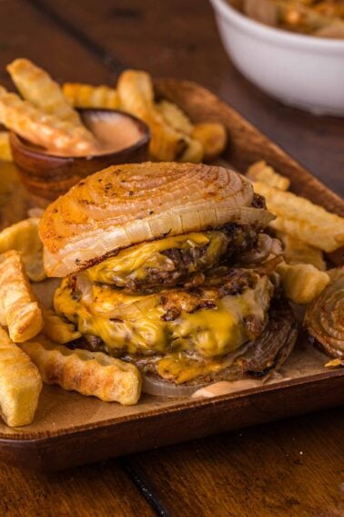 A Flying dutchman burger with grilled onions as buns, melted cheese, and crinkle-cut fries on a tray, with animal sauce on the side.