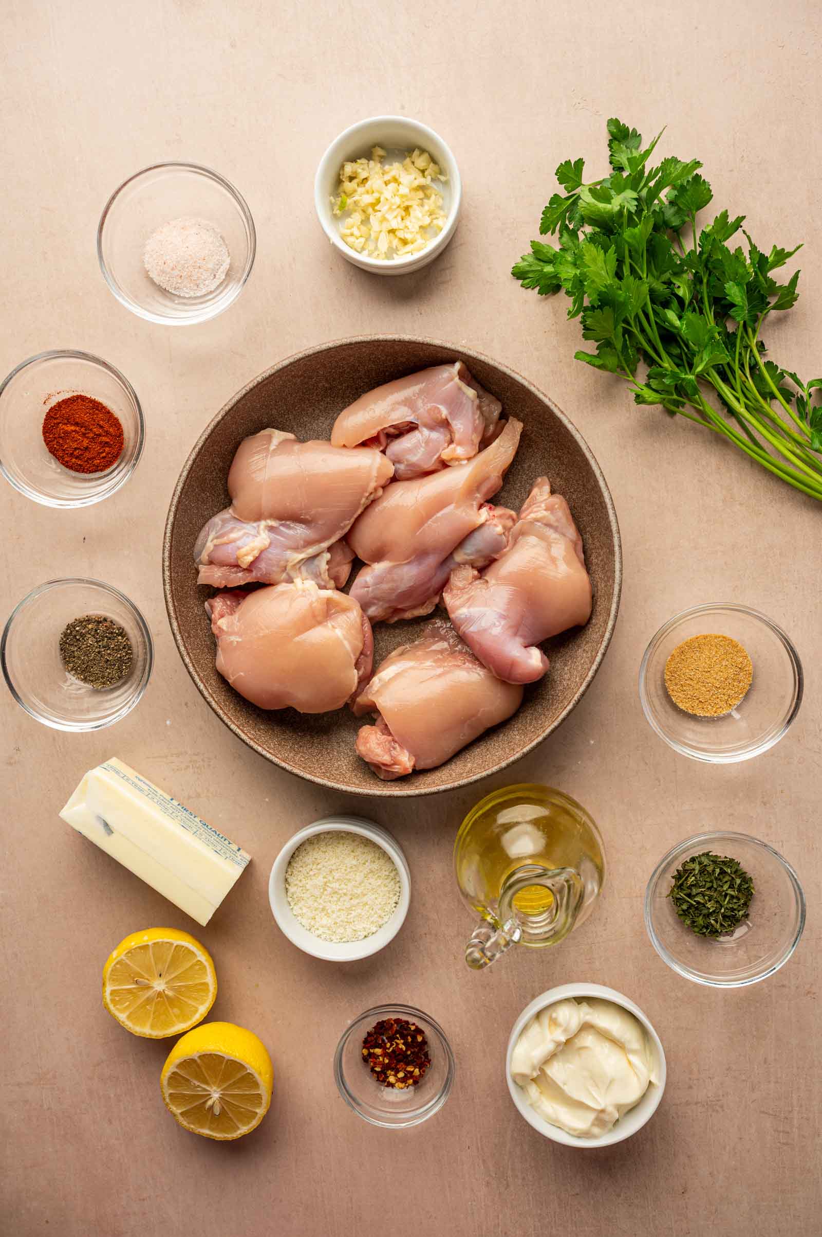 A bowl of raw chicken thighs surrounded by small bowls of spices, herbs, oil, butter, minced garlic, lemon halves, mayonnaise, and fresh parsley on a beige surface.