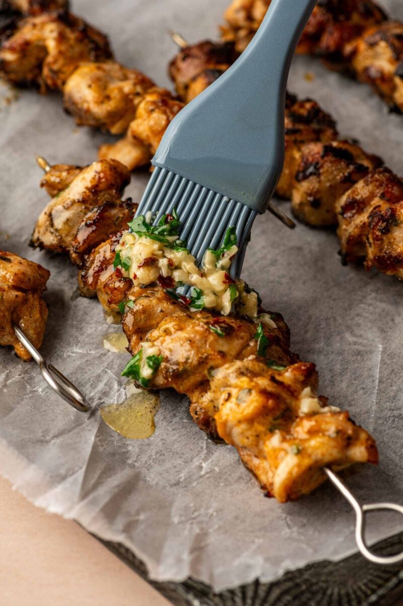 A silicone brush applies a garlic parmesan sauce to grilled chicken skewers resting on parchment paper.