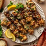 Four grilled chicken skewers garnished with herbs are served on a white plate with lemon wedges, garlicparm butter sauce, bread, garlic, and a green salad nearby.