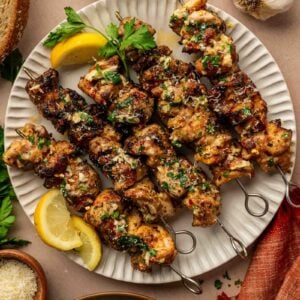 Four grilled chicken skewers garnished with herbs are served on a white plate with lemon wedges, garlicparm butter sauce, bread, garlic, and a green salad nearby.