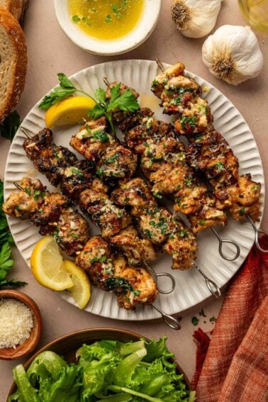 Four grilled chicken skewers garnished with herbs are served on a white plate with lemon wedges, garlicparm butter sauce, bread, garlic, and a green salad nearby.