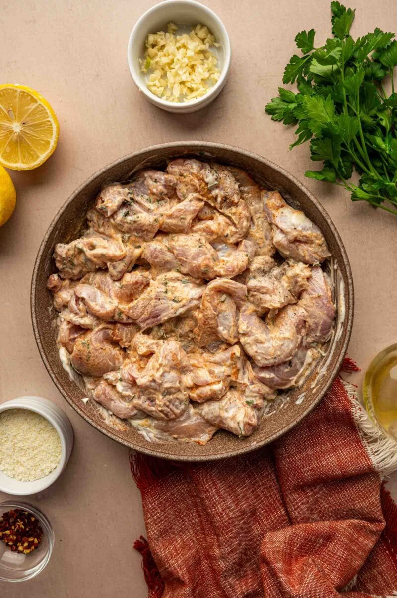 Raw marinated chicken pieces in a bowl, surrounded by chopped garlic, fresh parsley, lemon halves, red pepper flakes, and a jar of white powder on a beige surface with a red cloth.