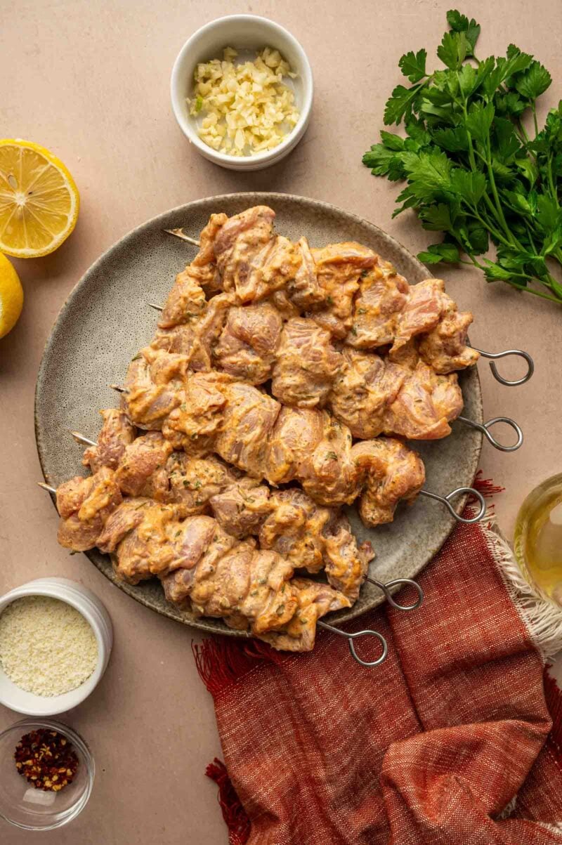 Raw marinated chicken skewers on a plate, surrounded by chopped garlic, parsley, lemon halves, grated cheese, and a red napkin on a beige surface.