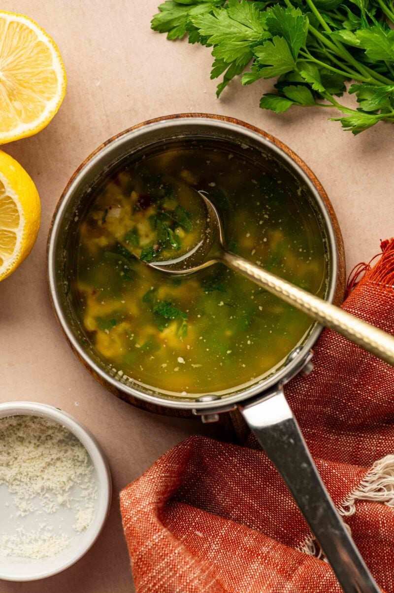 A saucepan filled with a garlic parmesan sauce, with a ladle inside. Fresh parsley, lemon halves, grated cheese, and a red cloth are nearby.
