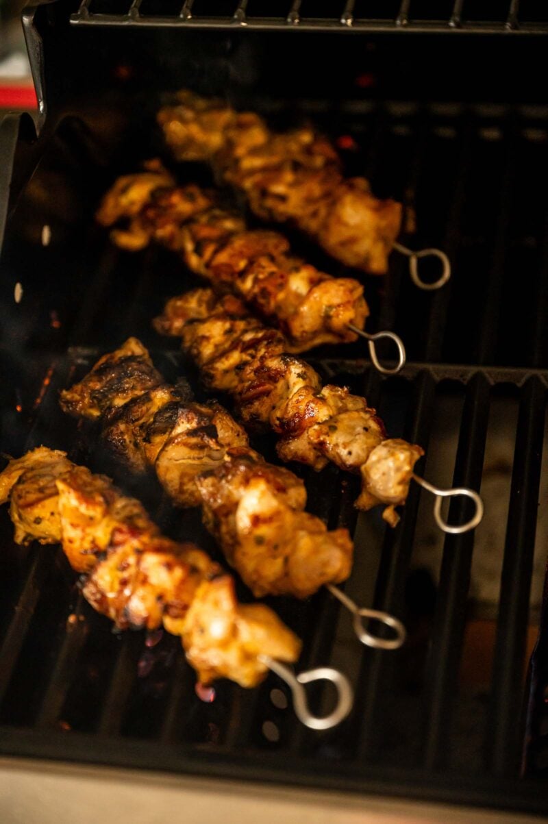Five skewers of marinated chicken are grilling on a barbecue, with visible grill marks and slight charring on the meat.