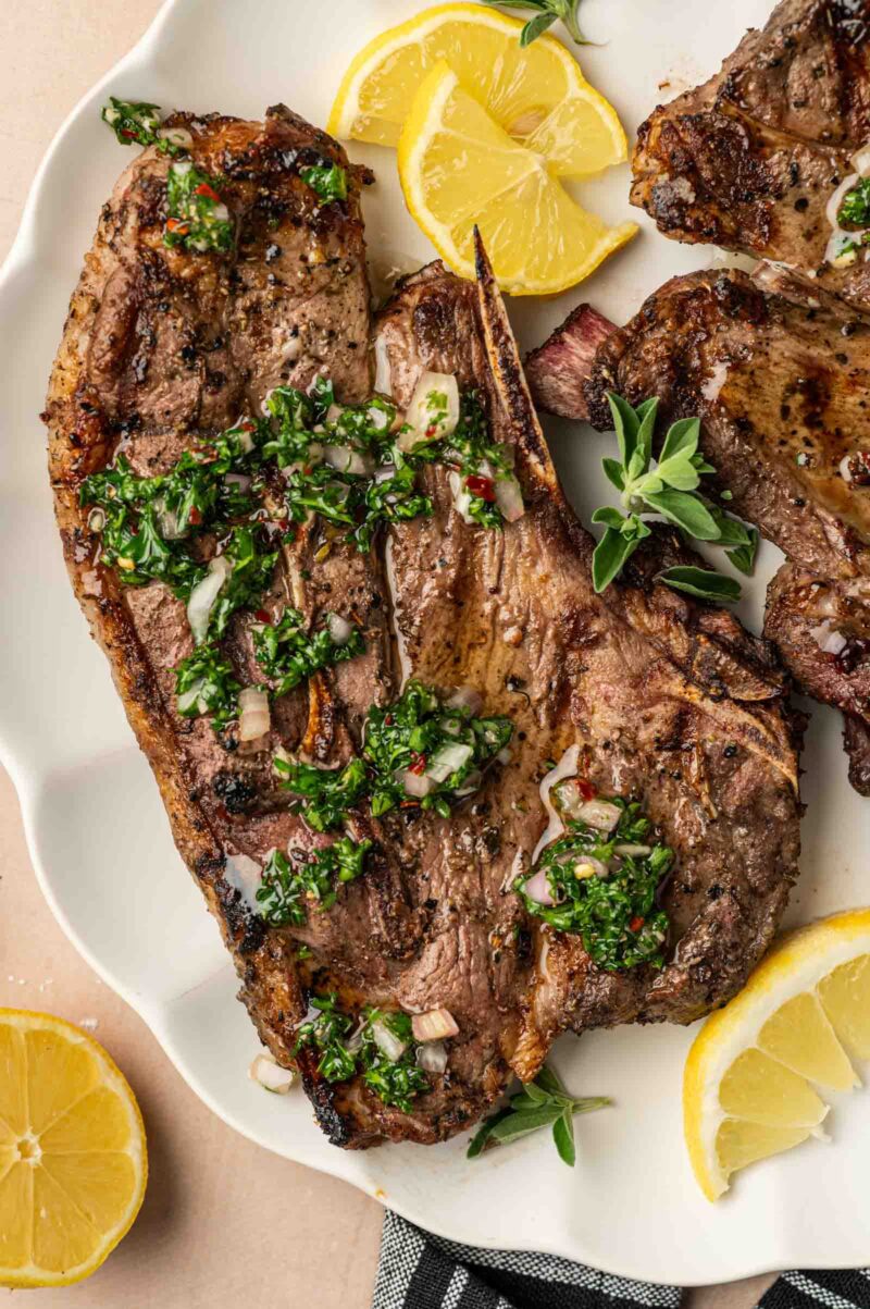 Grilled lamb shoulder chops topped with chimichurri sauce, garnished with fresh herbs and lemon wedges on a white plate.