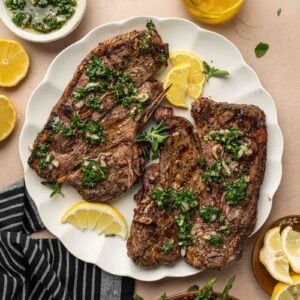Two grilled lamb shoulder chops topped with chimichurri sauce are served on a white plate with lemon slices, surrounded by grilled asparagus, a bowl of more sauce, lemon wedges, and a bottle of olive oil.