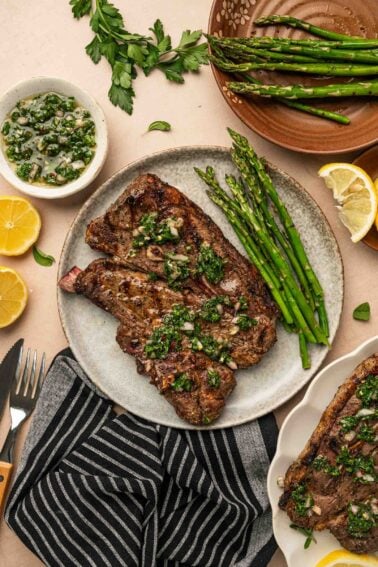 Grilled lamb shoulder chops topped with chimichurri sauce, served with asparagus spears on a plate, surrounded by lemon halves, herbs, and extra sauce on the table.