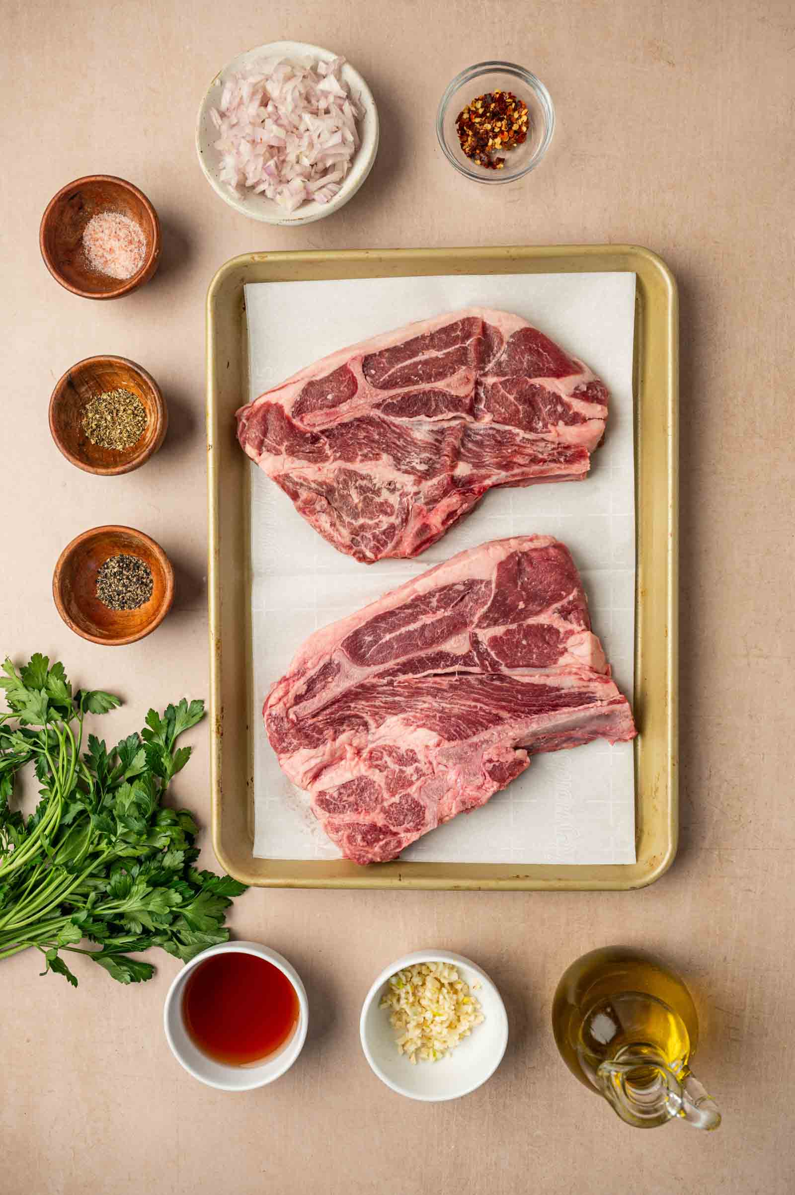 Two raw lamb shoulder chops on a tray with bowls of chopped shallots, red pepper flakes, salt, black pepper, parsley, olive oil, vinegar, and minced garlic arranged around them.