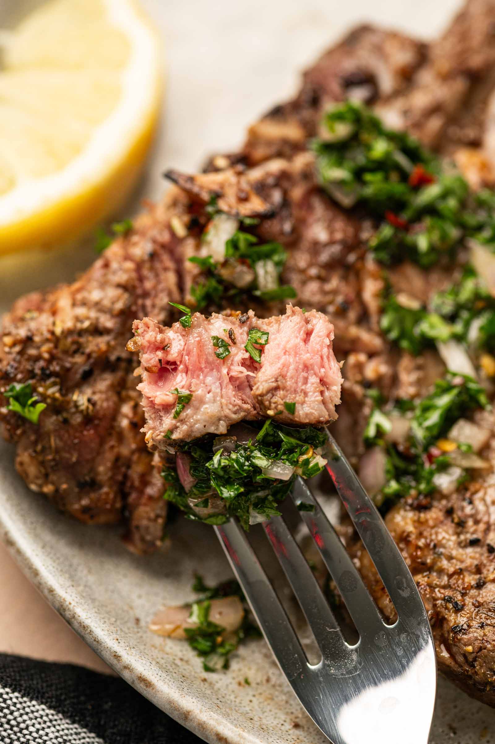 A close-up of a fork holding a piece of grilled stlamb shoulder chop topped with chopped herbs, with more and a lemon wedge on a plate in the background.