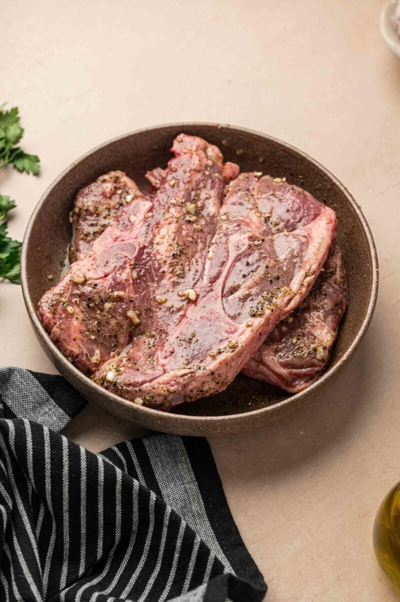 Raw seasoned lamb shoulder chops sit in a brown bowl on a beige surface, with a black and white striped cloth and parsley nearby.