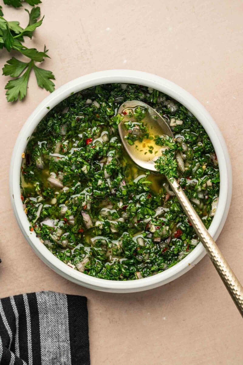 A white bowl of chimichurri sauce with chopped herbs and oil, with a spoon resting inside. Parsley leaves and a striped cloth are nearby on the table.