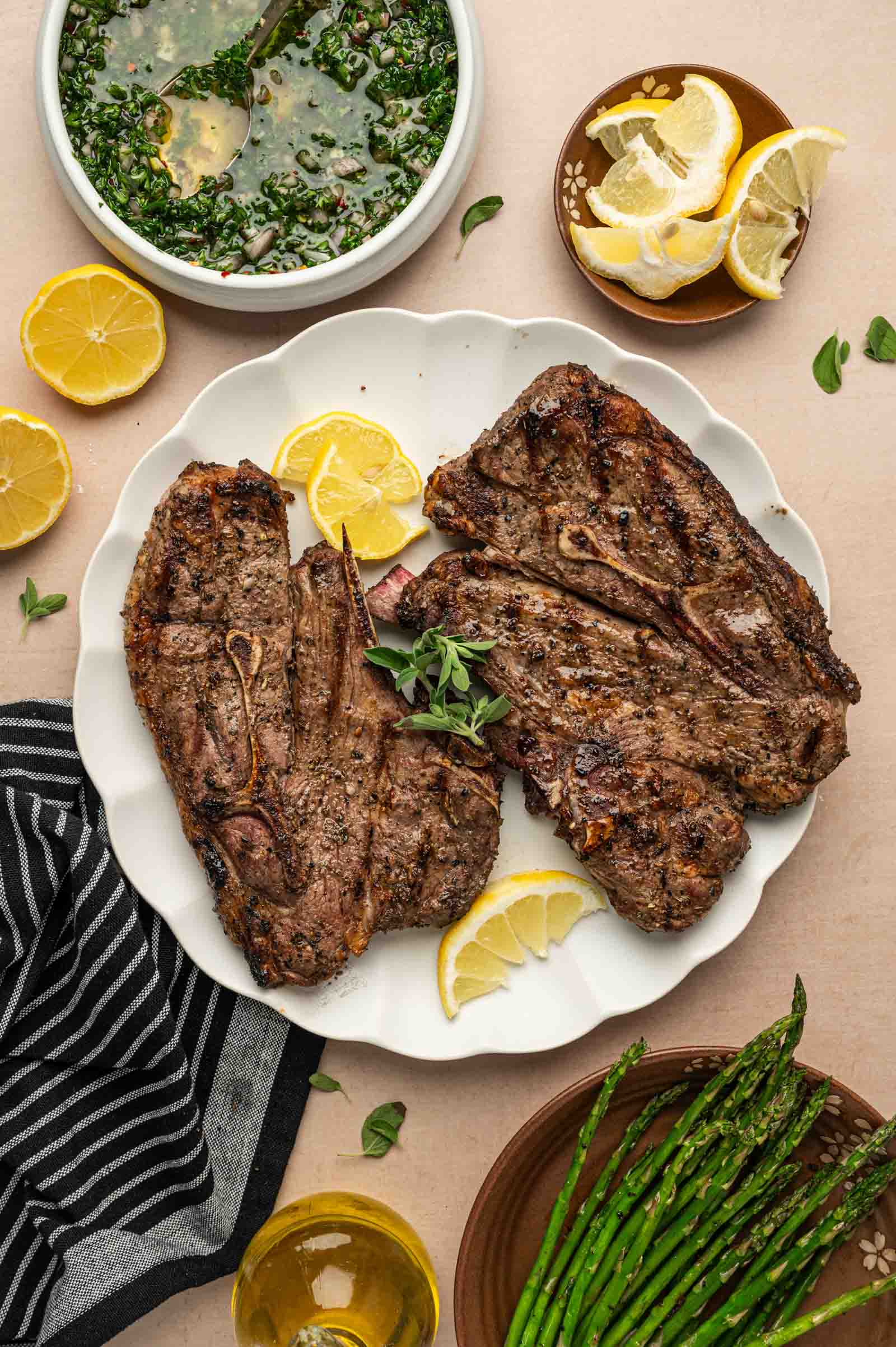 Three grilled lamb shoulder chops on a white plate with lemon wedges, served with grilled asparagus, a bowl of chimichurri sauce, lemon slices, and a striped napkin.