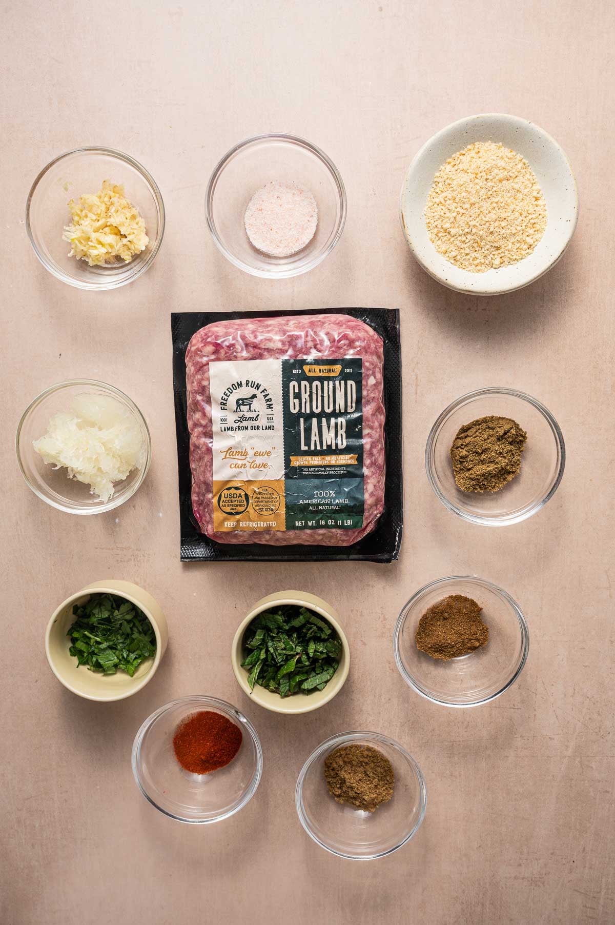 A package of ground lamb surrounded by small bowls holding various spices, herbs, garlic, grated onion, breadcrumbs, and salt, all arranged on a light surface.