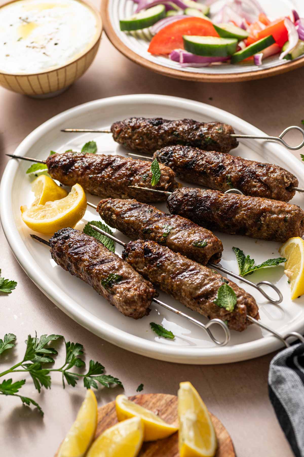 Grilled lamb koftas on metal skewers served on a white plate with lemon wedges and mint leaves; salad and sauce bowls are in the background.