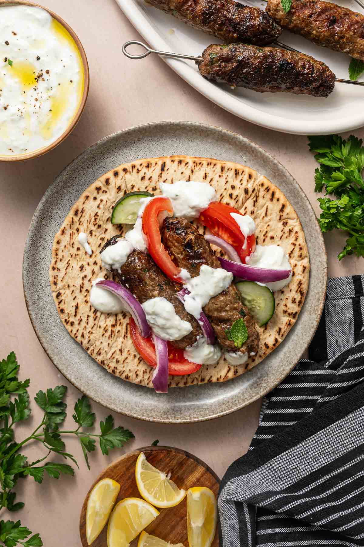 A plate with pita bread topped with grilled lamb kofta, sliced tomatoes, cucumber, red onions, and tzatziki sauce. Surrounding the plate are herbs, lemon wedges, and more koftas.