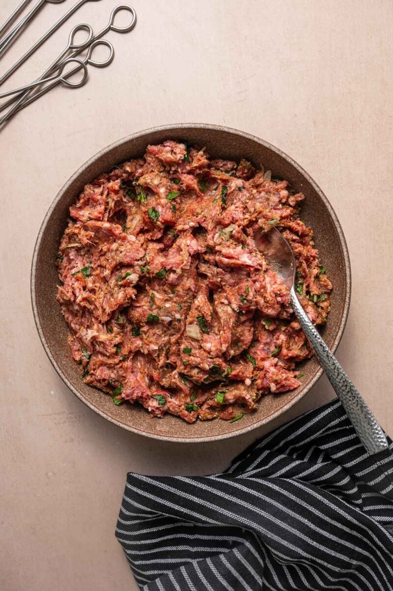 A bowl of seasoned ground lamb mixture with herbs, a spoon inside, metal skewers beside, and a striped cloth at the bottom edge.