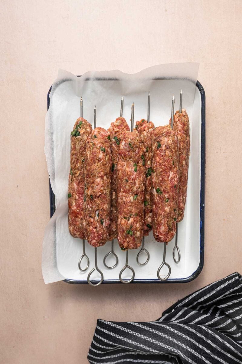 Raw ground lamb koftas on metal skewers rest in a white tray lined with parchment paper, placed on a light surface next to a black and white striped cloth.