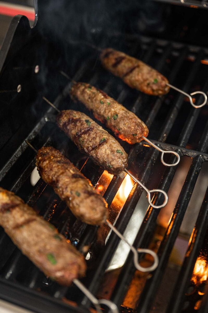 Five lamb kofta skewers are being grilled over an open flame on a barbecue grill, with visible smoke and grill marks on the meat.