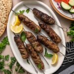 Grilled lamb kofta on metal skewers served on a white oval plate with lemon wedges and mint leaves, surrounded by pita bread, tzatziki sauce, and a cucumber-tomato salad.