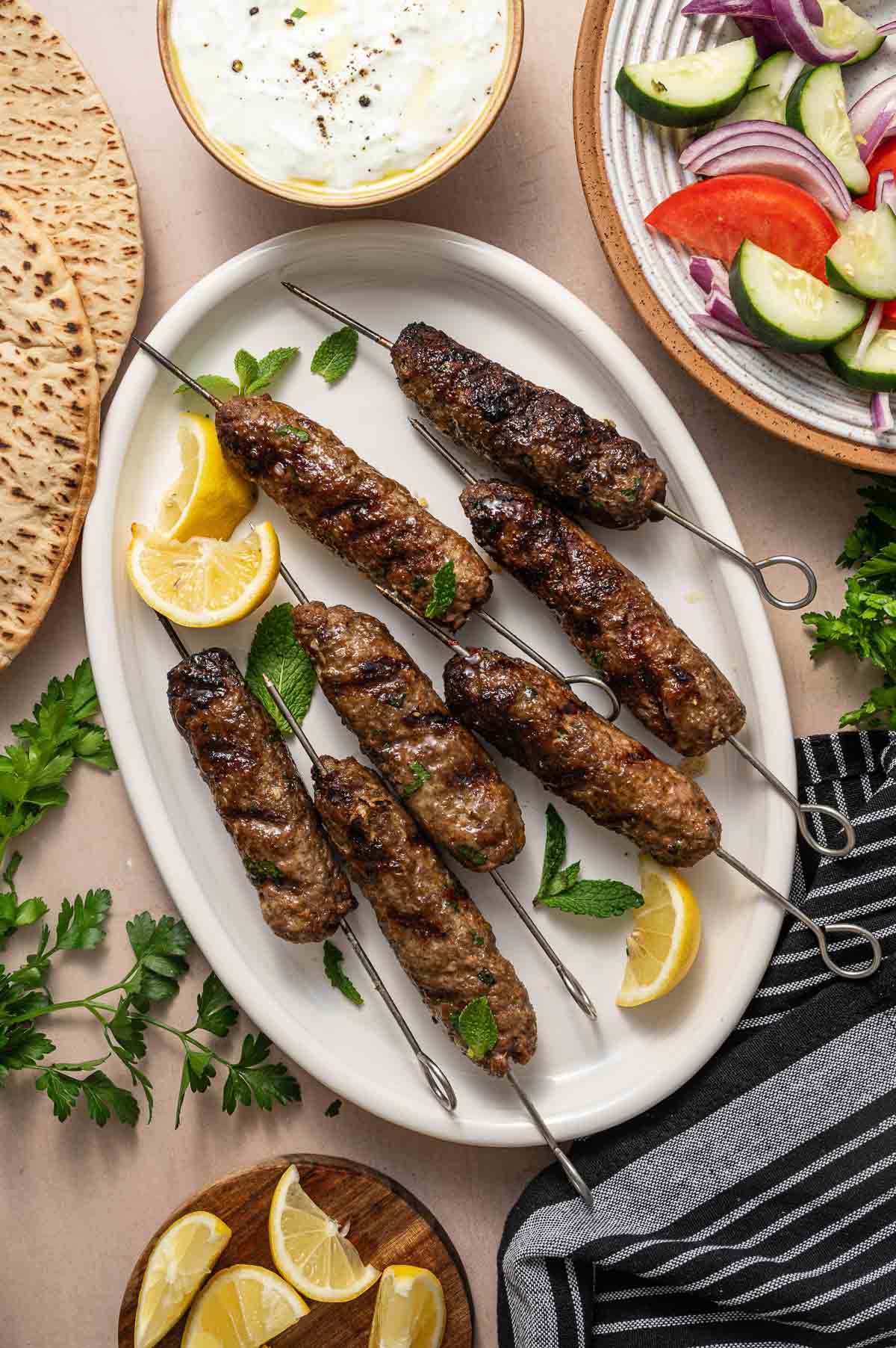 Grilled lamb kofta on metal skewers served on a white oval plate with lemon wedges and mint leaves, surrounded by pita bread, tzatziki sauce, and a cucumber-tomato salad.