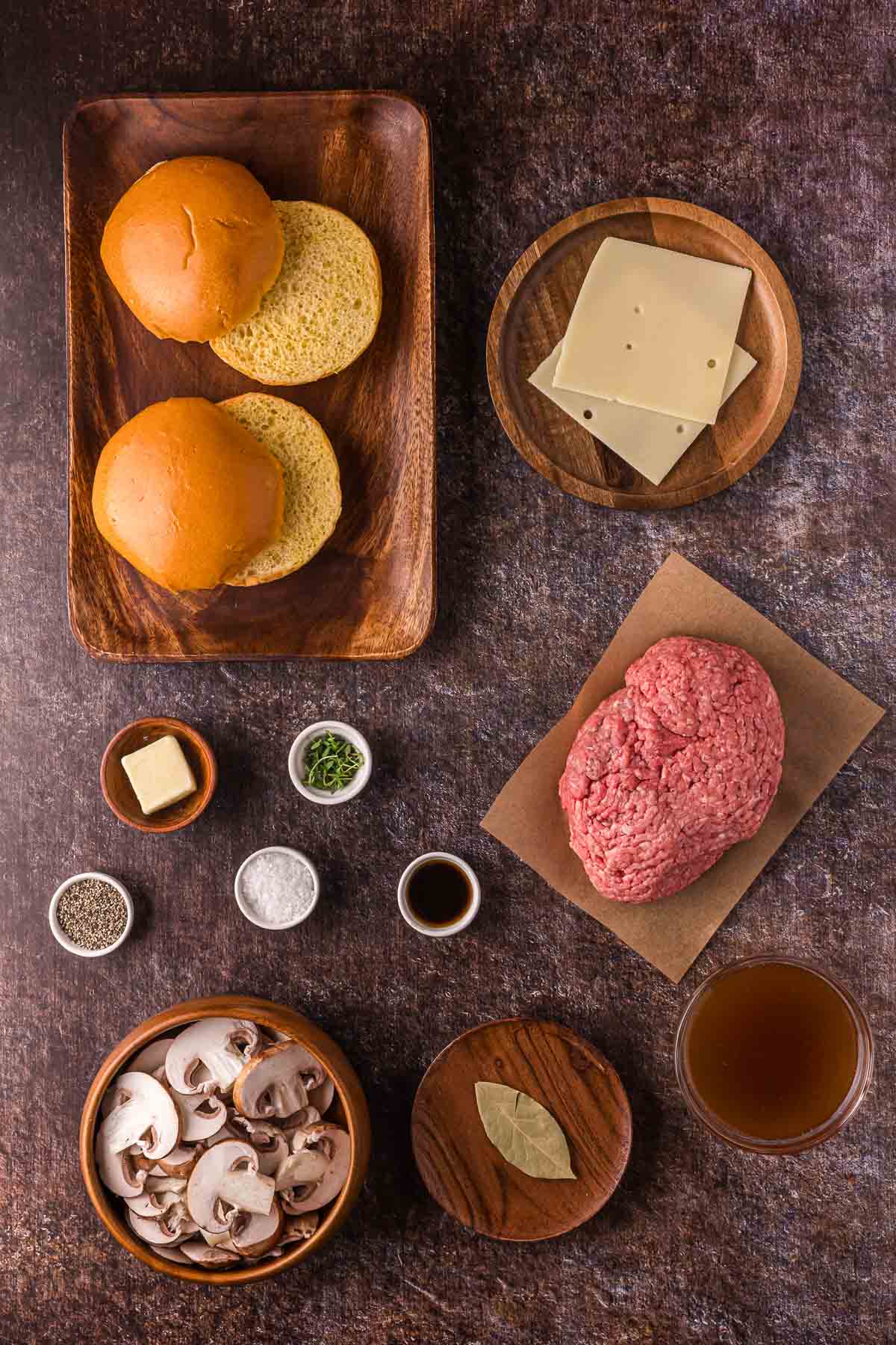 Top-down view of burger buns, Swiss cheese slices, ground beef, sliced mushrooms, butter, seasonings, Worcestershire sauce, a bay leaf, and beef broth arranged on a dark surface.