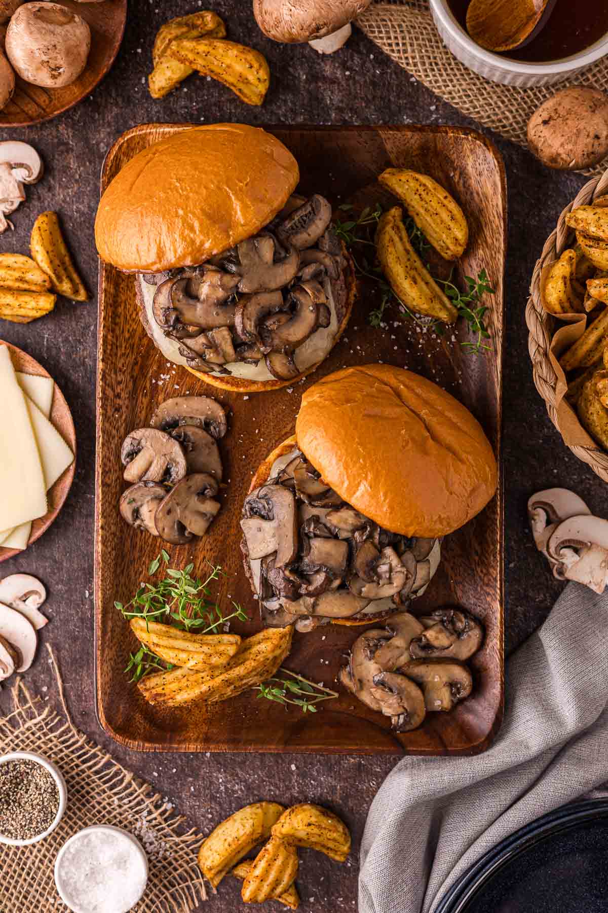 Two mushroom Swiss burgers on a wooden tray with potato wedges, surrounded by sliced mushrooms, cheese slices, herbs, and condiments on a rustic table.