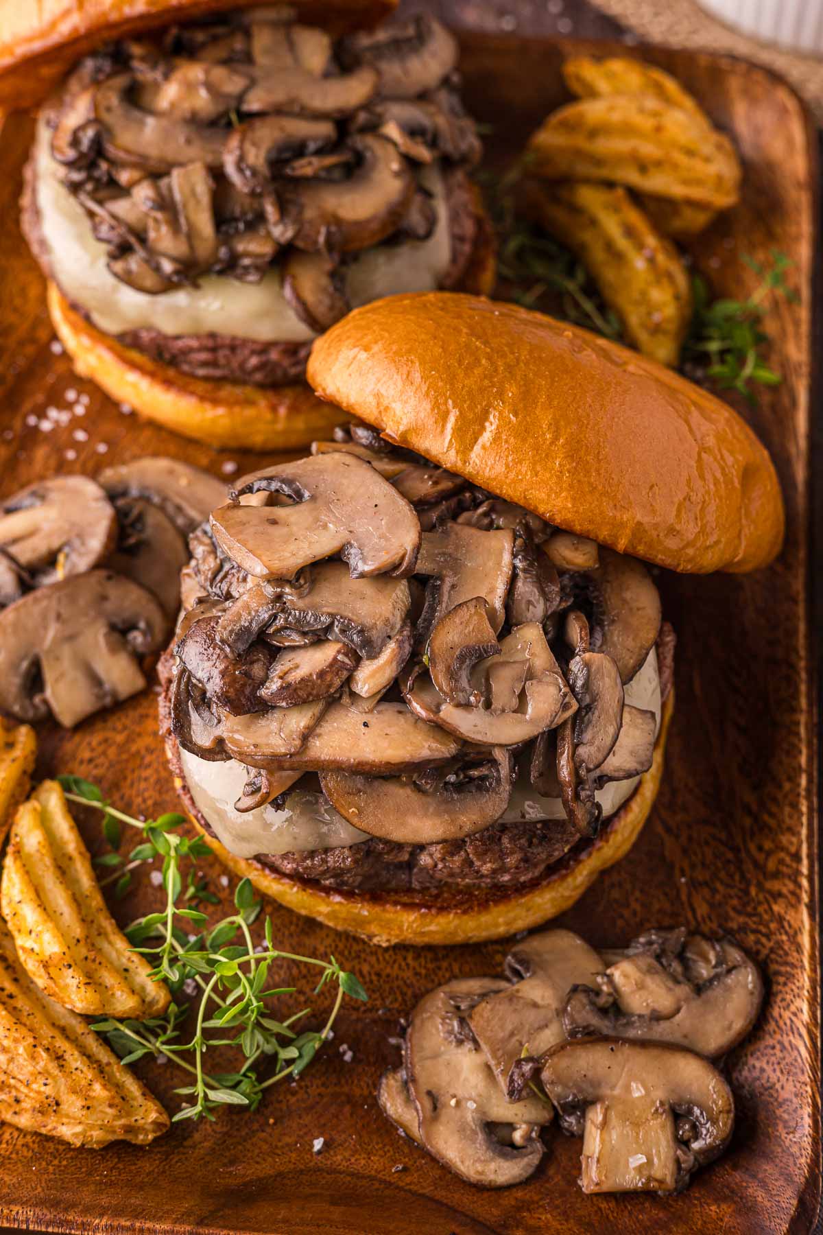 A burger topped with sautéed mushrooms and melted swiss cheese on a wooden tray, served with waffle fries and garnished with fresh herbs.