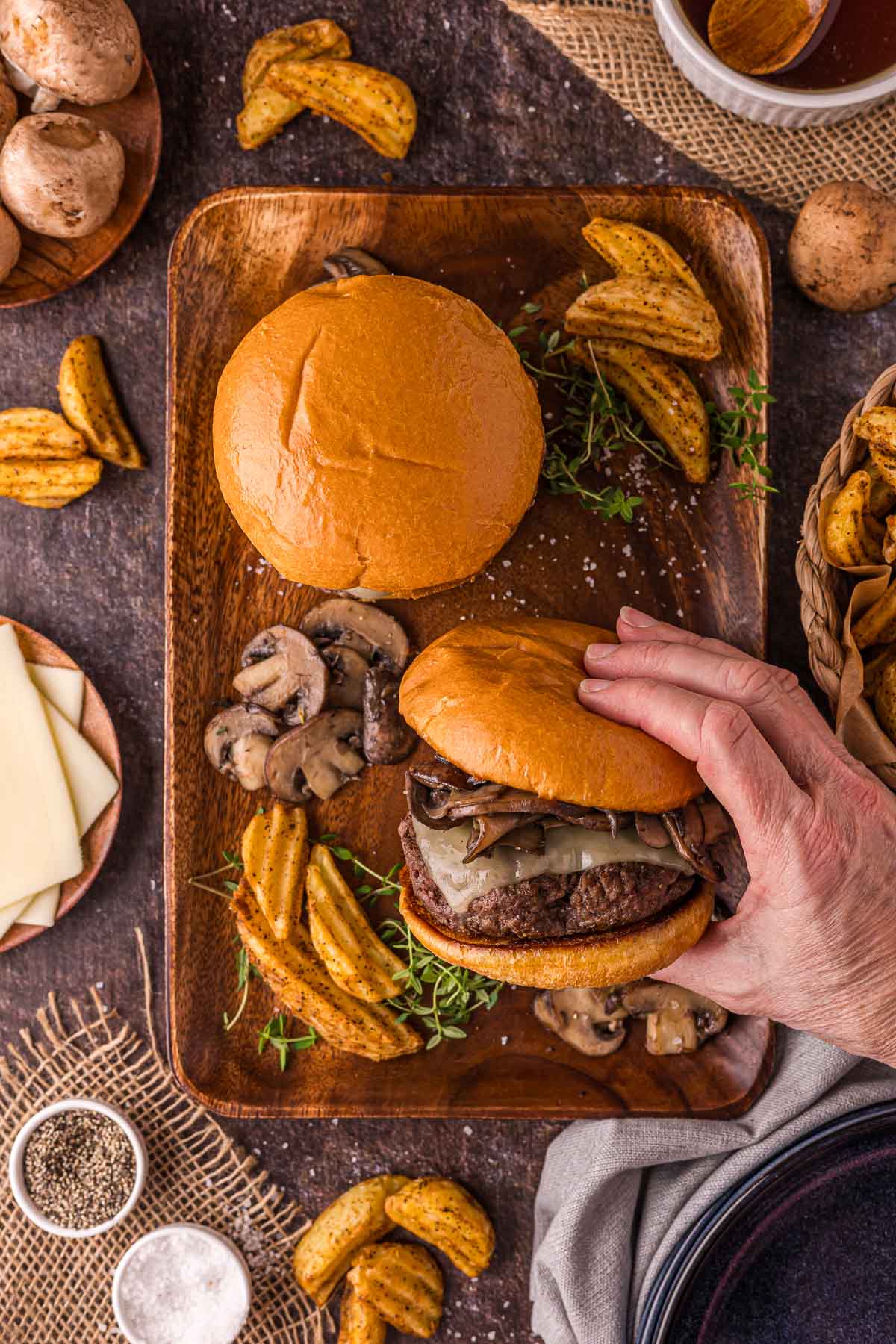 A hand holds a mushroom swiss burger wover a wooden tray, surrounded by potato wedges and burger bun.