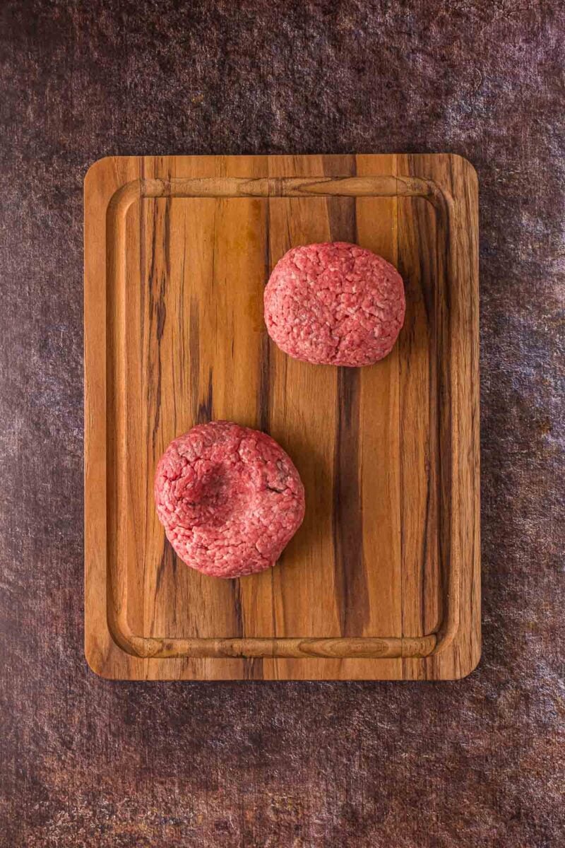 Two raw ground beef patties placed on a wooden cutting board, set against a textured dark surface.