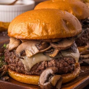 A hamburger topped with melted cheese and sautéed mushrooms on a wooden plate, with fries and a bowl in the background.