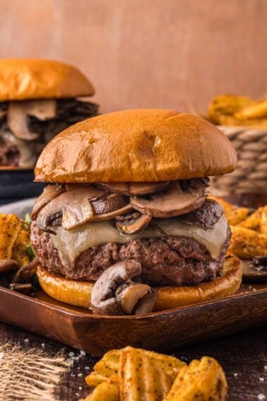 A cheeseburger with sautéed mushrooms on a brioche bun is served with crinkle-cut fries on a wooden plate.