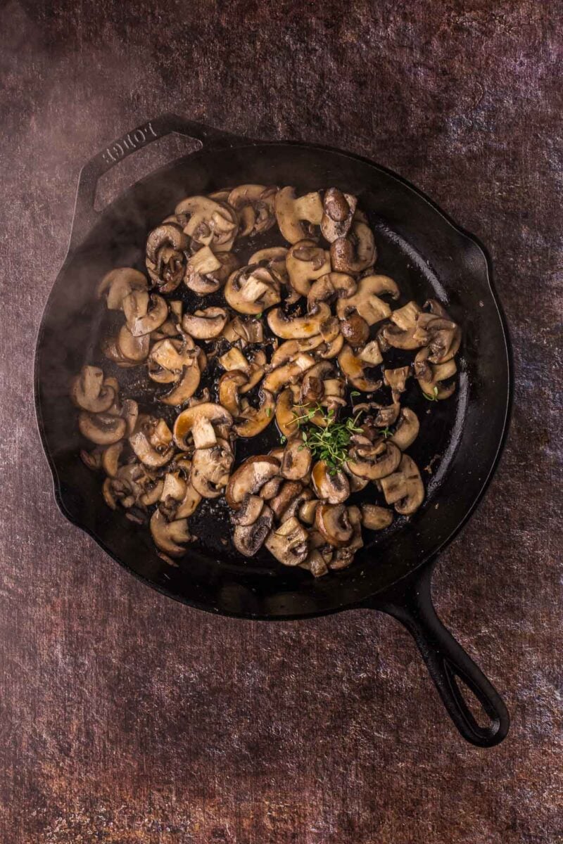 Sliced mushrooms sautéing in a black cast iron skillet with fresh herbs on a textured brown surface.