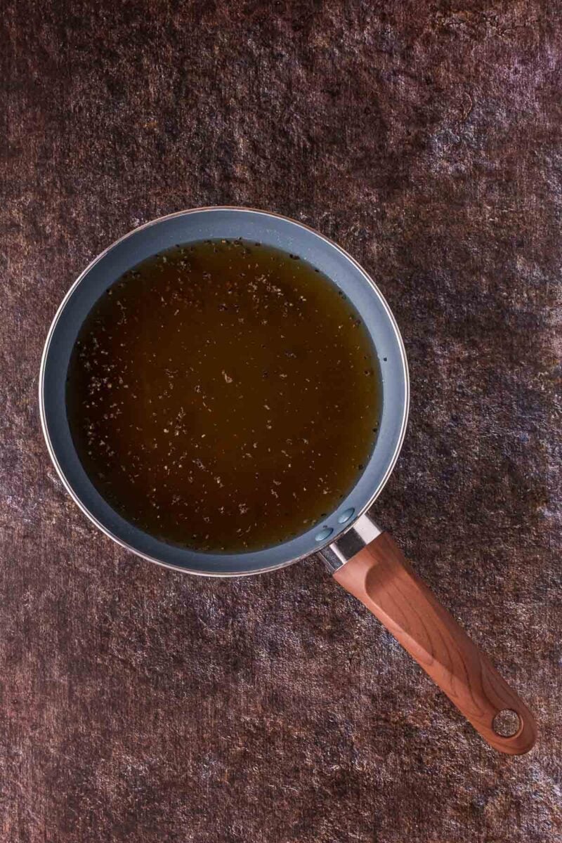 A frying pan filled with au jus sits on a textured brown surface.