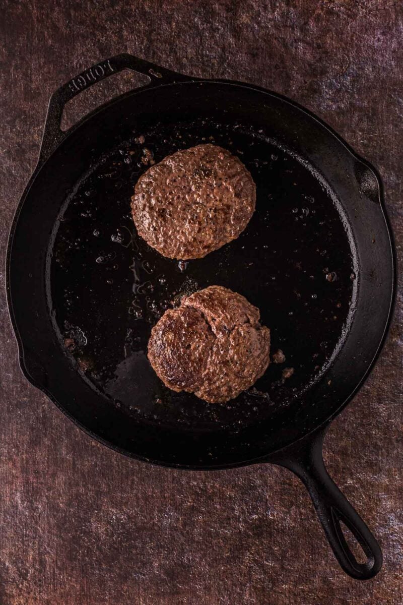 Two cooked burger patties sit in a black cast-iron skillet on a dark textured surface.