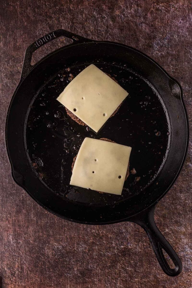 Two hamburger patties topped with slices of Swiss cheese are cooking in a black cast iron skillet.