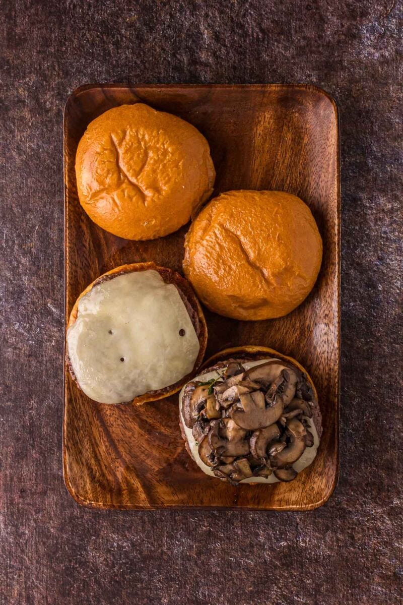 A wooden tray with two closed burger buns and one open burger showing melted cheese and sautéed mushrooms on a beef patty.