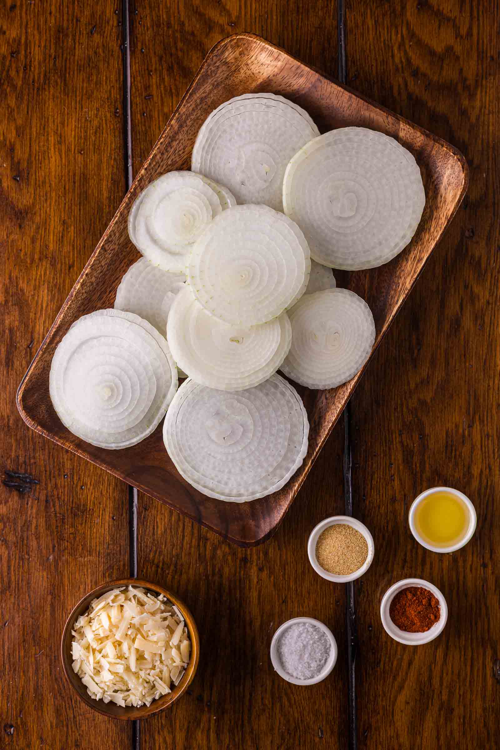 A wooden tray with sliced yellow onions, next to small bowls of shredded cheese, oil, salt, paprika, and onion powder on a wooden table.