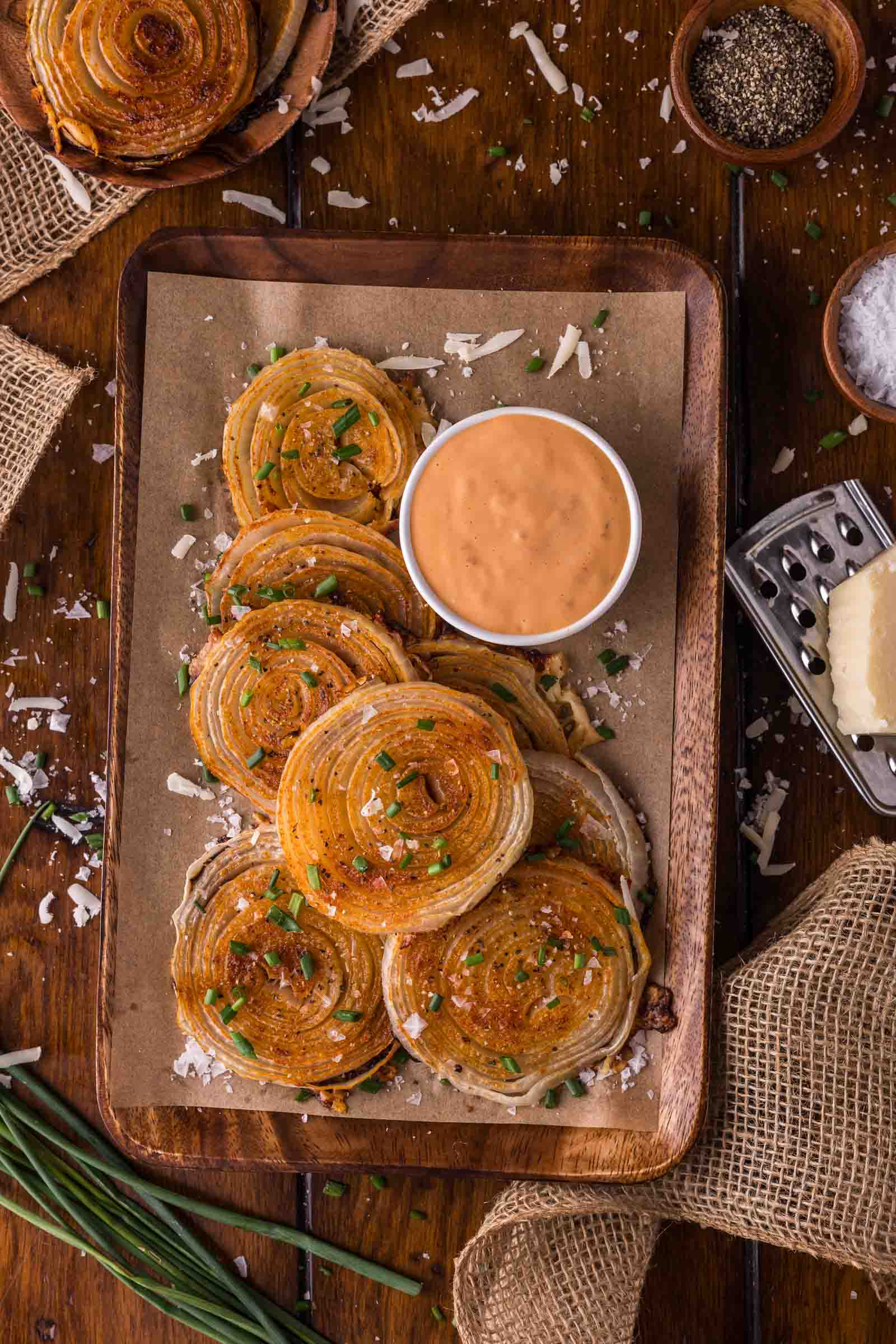 A wooden tray with onion chips garnished with herbs, served with a small bowl of animal sauce, grated cheese, and seasonings on the side.