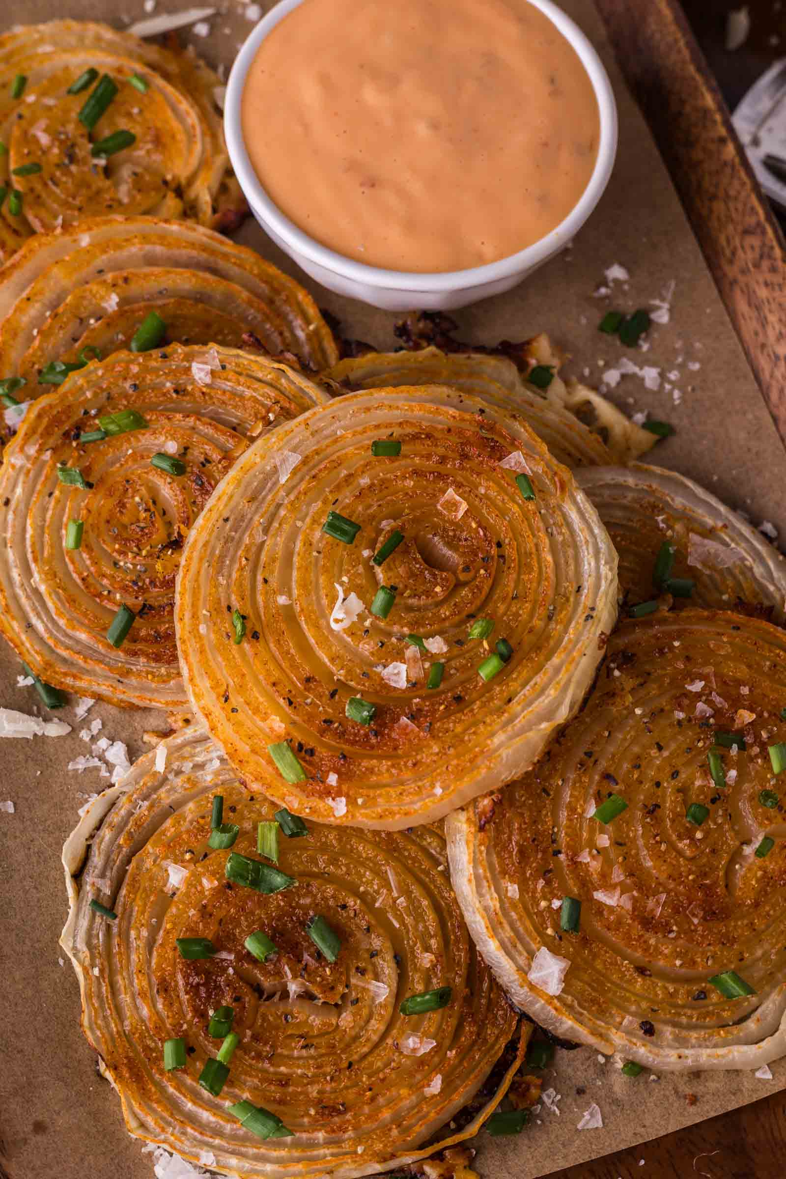 Sliced, roasted onion chips garnished with chopped chives and salt, served on parchment paper with a bowl of creamy dipping sauce.