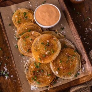 A wooden tray with crispy roasted onion chips topped with chopped chives and coarse salt, served with a bowl of creamy dipping sauce.